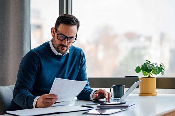 A middle aged man in glasses and a white collar shirt, underneath a blue sweater, is looking of some papers and working on his computer