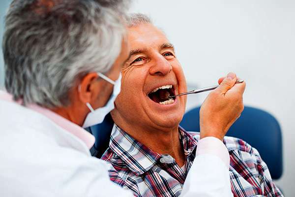 A gray-haired dentist seated next to him holds an examination tool in the patient's mouth. The patient is wearing a plaid shirt, and the dentist is wearing a white coat and mask.