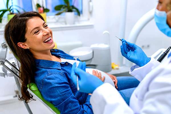 Pregnant woman in a jean jacket smiling as her dentist prepares to examine her teeth.