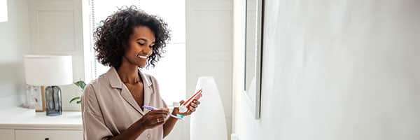 A woman  with dark, wavy hair, just above shoulder length, examining toothpaste while hold her purple toothbrush in her other hand